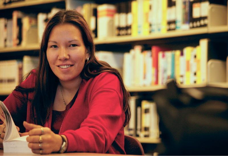 Woman holding an open book seated at a table in front of a bookshelf